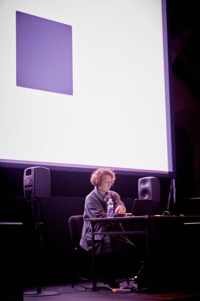 Person playing music from a laptop sitting in front of a screen of abstract projections.