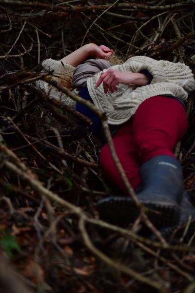 Person wearing red trousers and a white cardigan lying on the ground in a forest.