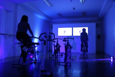 Two people watching TVs on exercise bikes in a dark studio